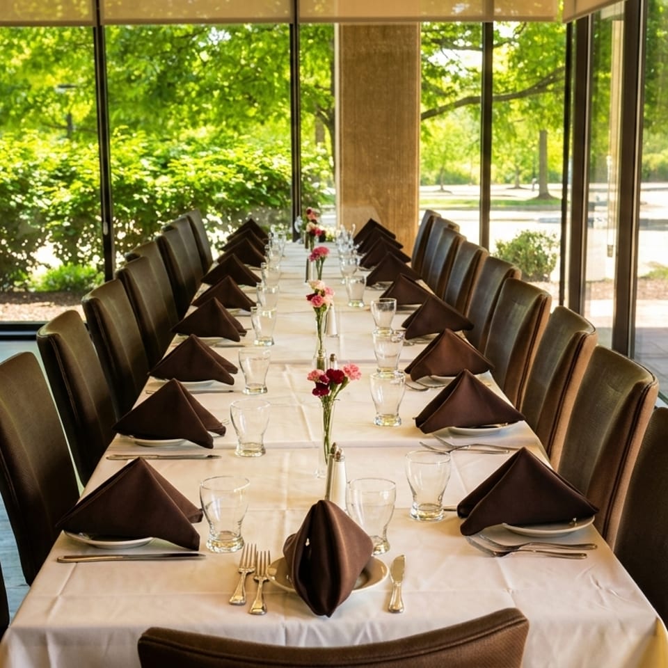 Long dining table set with white tablecloths, brown napkins, and small floral centerpieces, next to large windows.