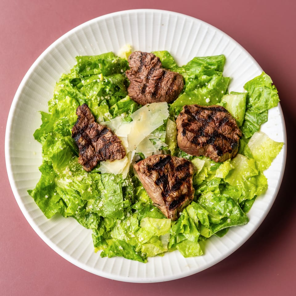 Grilled steak pieces top a Caesar salad with romaine lettuce, creamy dressing, and shaved Parmesan in a white bowl.