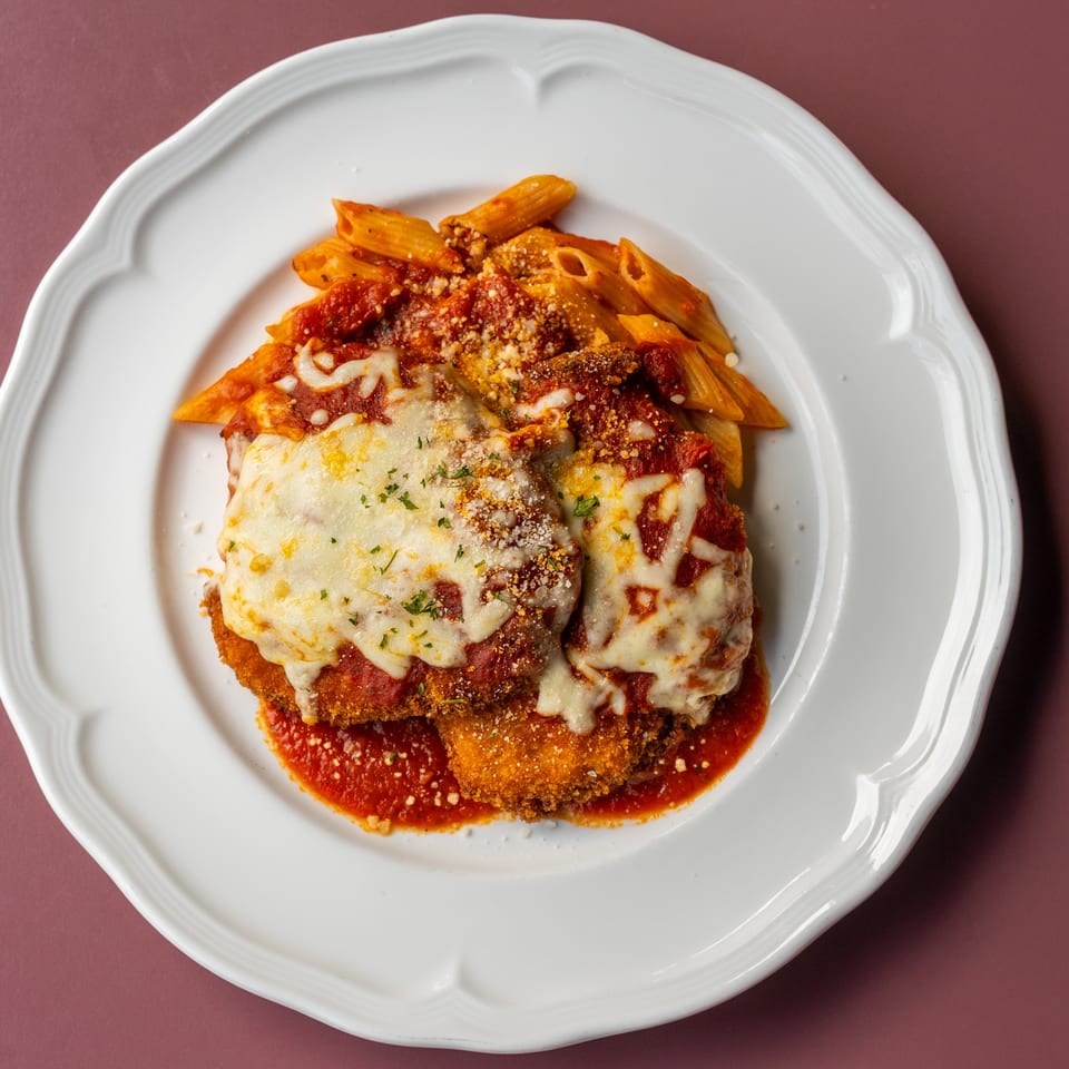 Breaded chicken Parmesan topped with melted cheese and marinara, served with penne pasta on a white plate.