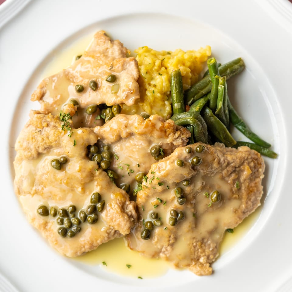 Breaded chicken cutlets with creamy caper sauce, yellow rice, and green beans on a white plate.