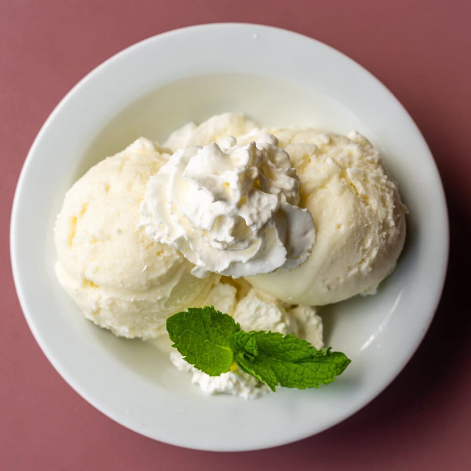 Scoops of vanilla ice cream topped with whipped cream and fresh mint in a white bowl on a red table.