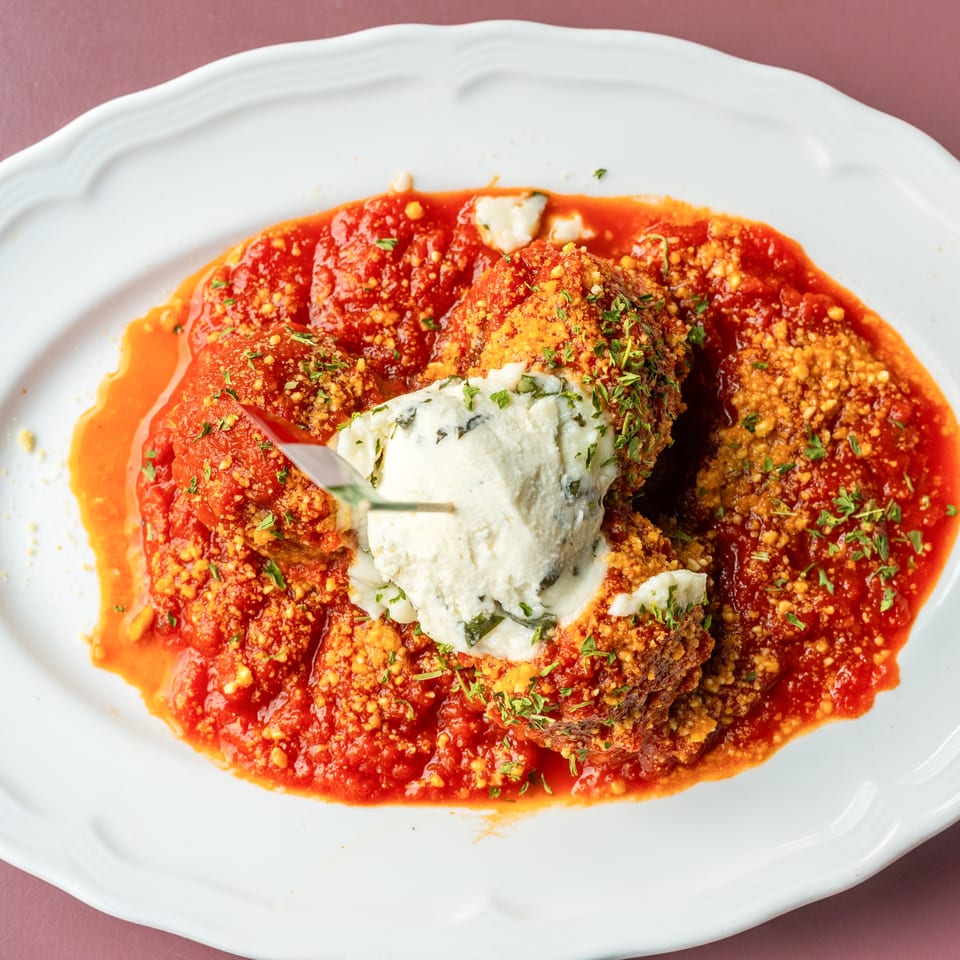 Crispy breaded meatballs topped with melted blue cheese and herbs, served in vibrant tomato sauce on a white platter.