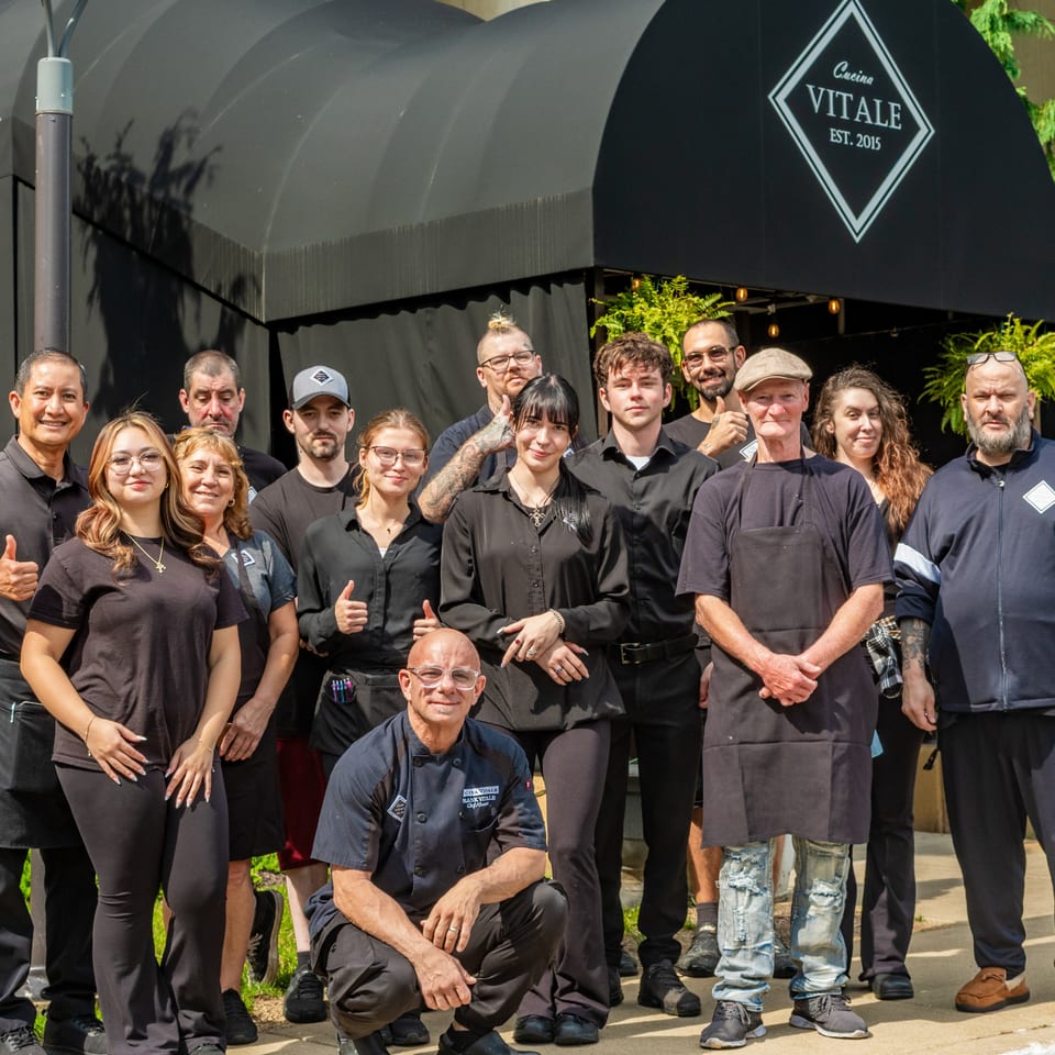 Cucina Vitale restaurant staff pose outside the entrance beneath a black awning bearing the restaurant’s logo.