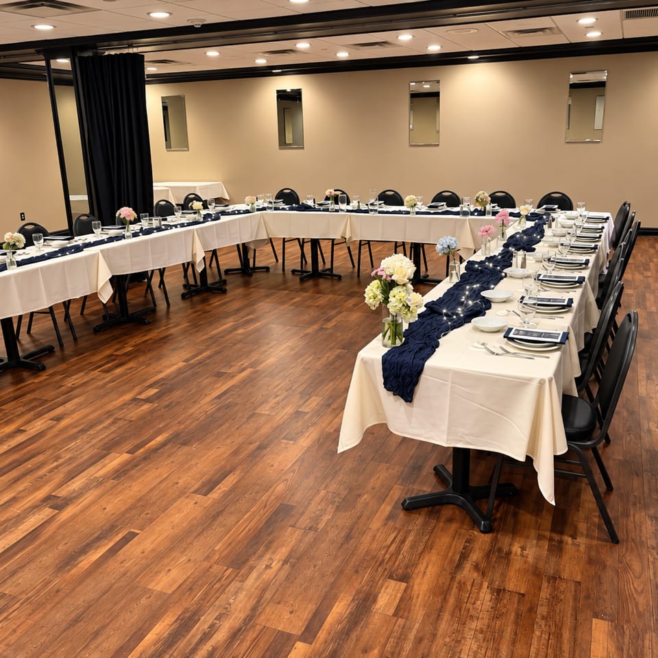 Elegant banquet room with U-shaped tables, white linens, navy runners, floral centerpieces, place settings, and polished wood floors.