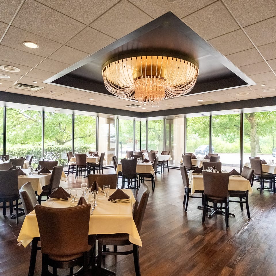 Elegant restaurant dining room with white tablecloths, dark chairs, floor-to-ceiling windows, and a large illuminated chandelier.