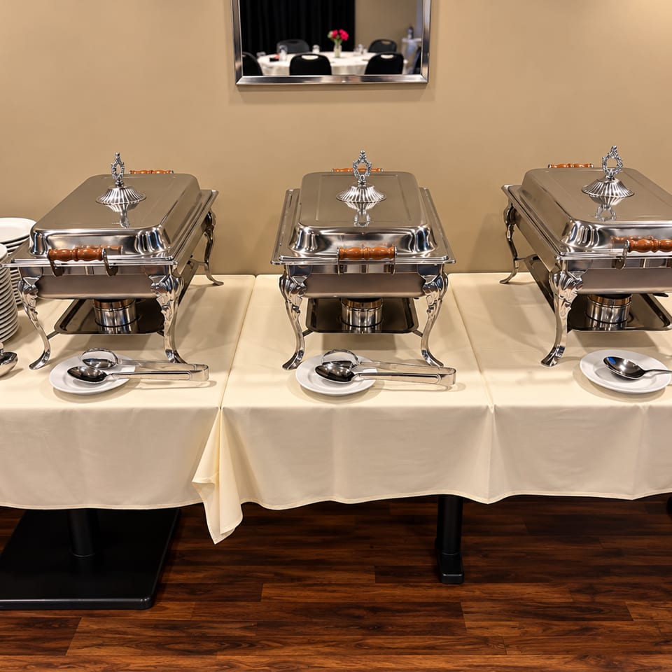 Three polished silver chafing dishes sit on a white buffet table with stacked plates, serving utensils, and a dining room mirror.