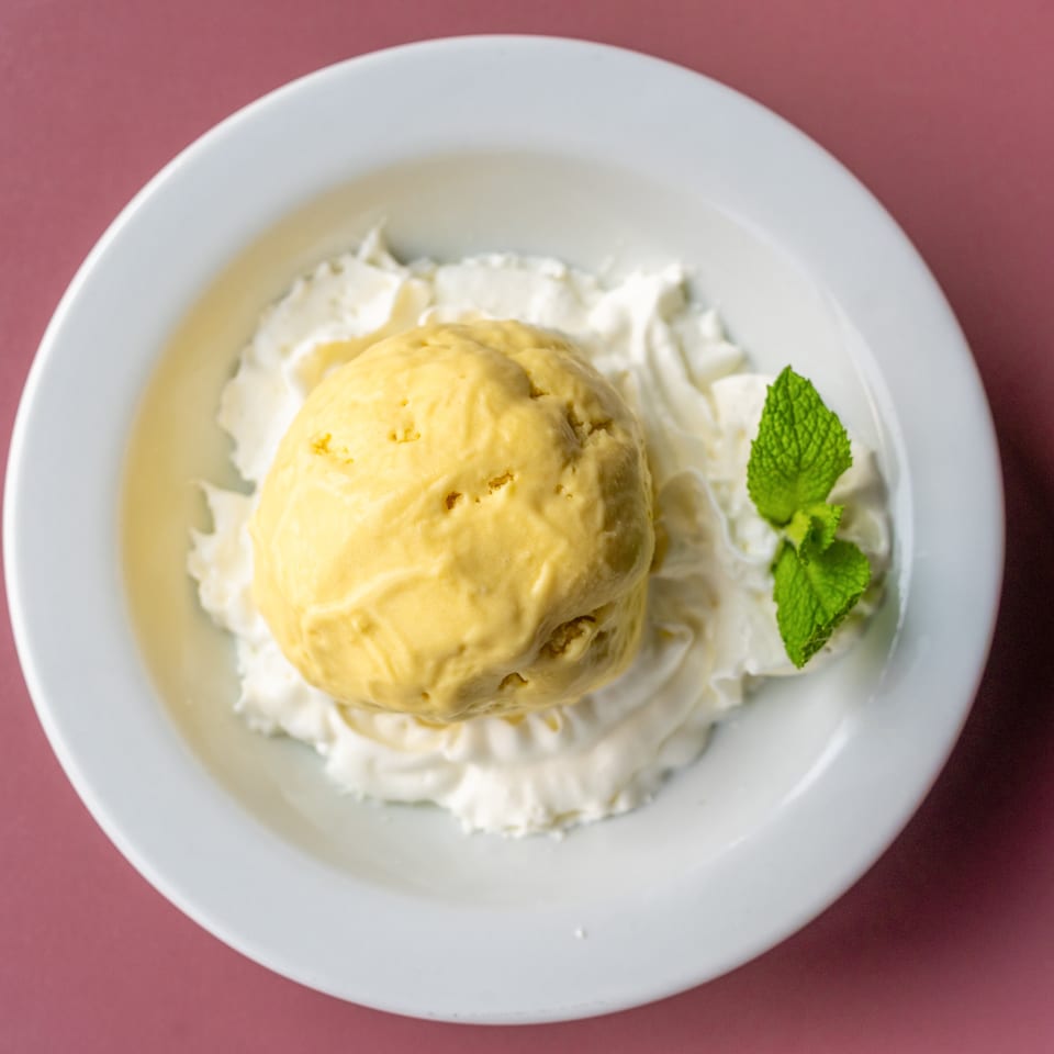 Scoop of yellow ice cream surrounded by whipped cream and garnished with fresh mint on a white plate.