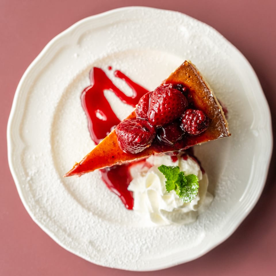 Slice of berry-topped cheesecake with whipped cream, raspberry sauce, and mint garnish on a white plate.