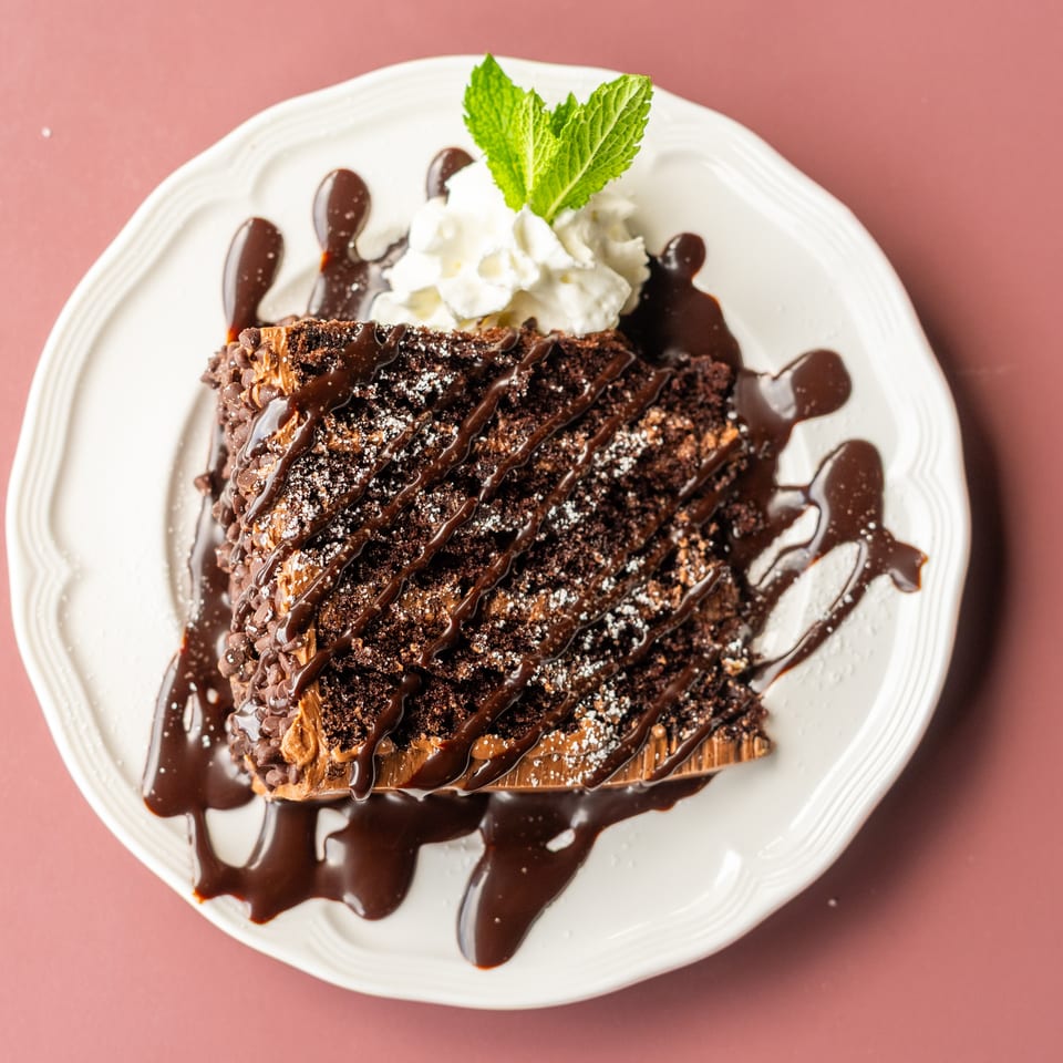 Chocolate cake slice drizzled with chocolate sauce, powdered sugar, and whipped cream, garnished with mint on a white plate.