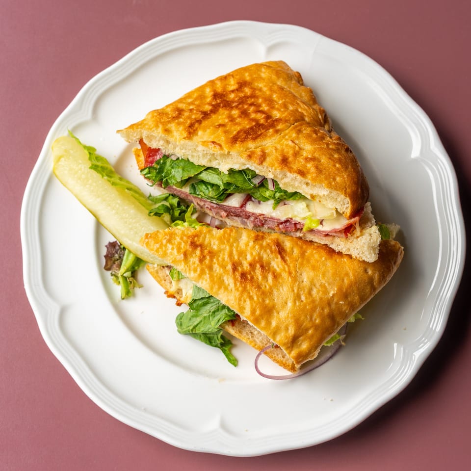 Toasted deli sandwiches filled with meats, cheese, lettuce, and vegetables, served with a pickle spear on a white plate.
