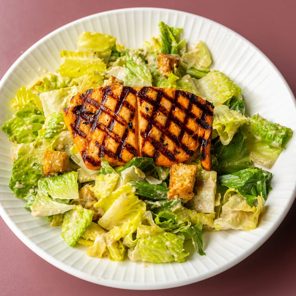 Grilled chicken breast served over creamy Caesar salad with romaine lettuce and croutons on a white plate.
