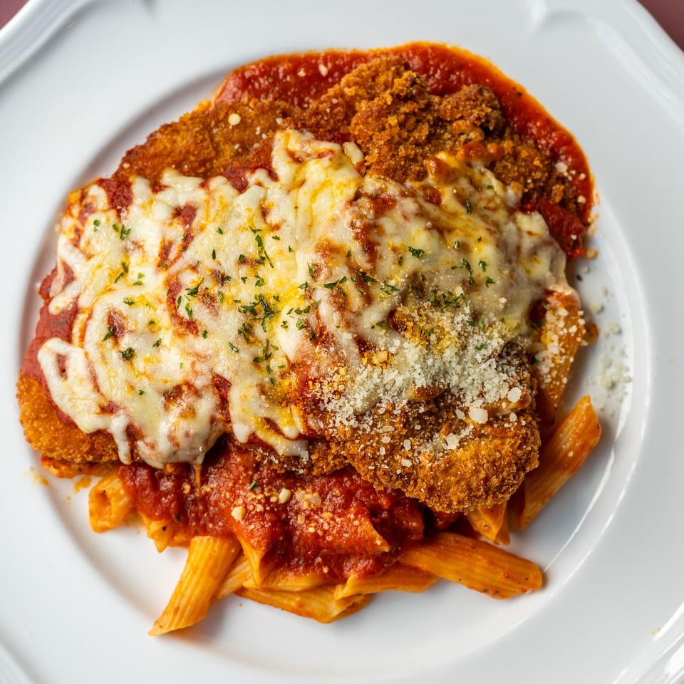 Breaded chicken Parmesan topped with melted cheese and herbs, served over penne pasta in tomato sauce.