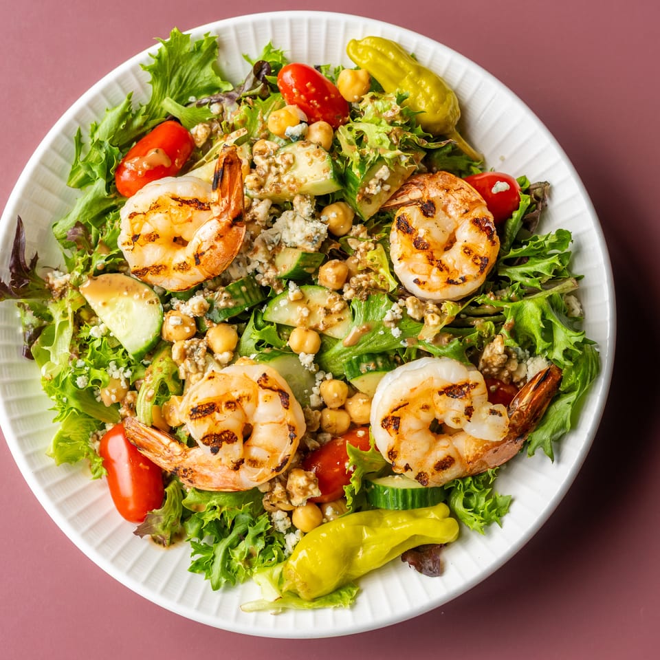 Grilled shrimp salad with mixed greens, cucumbers, tomatoes, chickpeas, blue cheese, dressing, and pepperoncini in a white bowl.