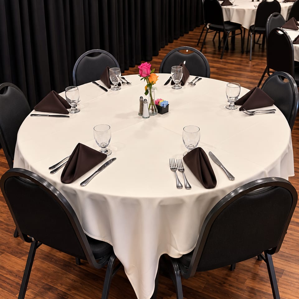 Round banquet table set with white linens, brown napkins, glassware, silverware, and a floral centerpiece in a wood-floored dining room.