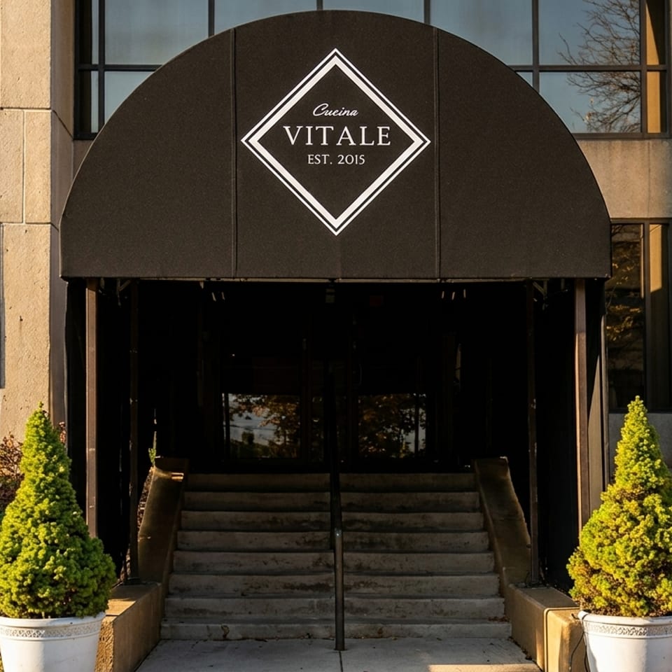 Cucina Vitale restaurant entrance with a black awning, concrete steps, and potted evergreen plants.